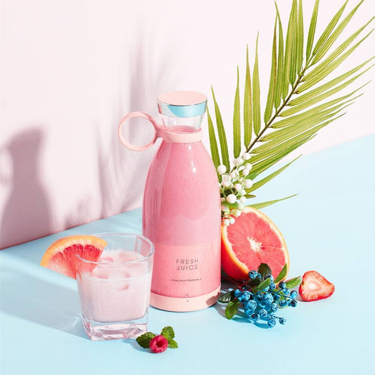 Portable Blendy Bottle blender with pink design next to a glass of juice, grapefruit, berries, and a palm leaf.